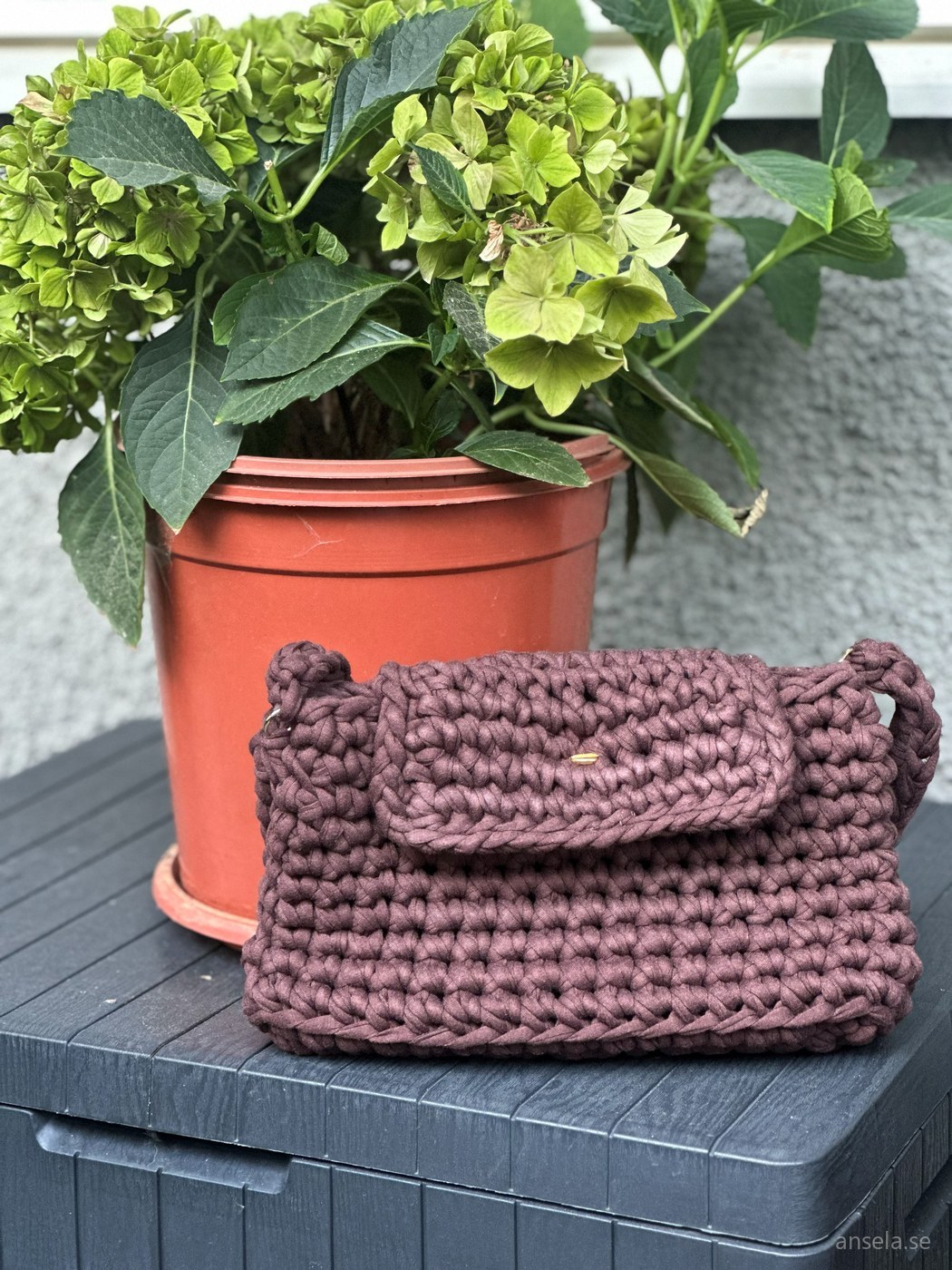 Virkad väska i dovt rosenbrunt på ett utebord bredvid en kruka med hortensia
